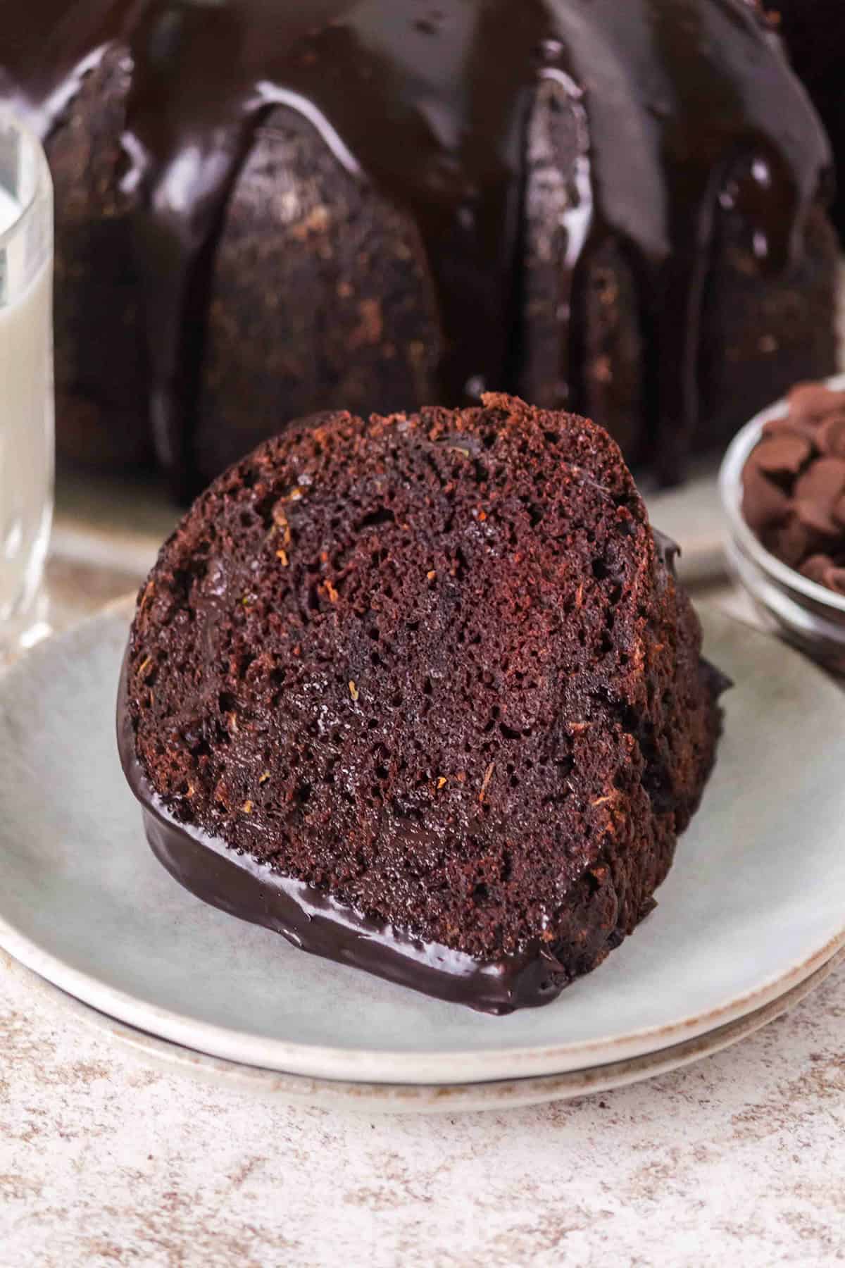 A slice of Triple Chocolate Zucchini Bundt Cake with rich chocolate glaze sits on a plate, accompanied by a glass of milk and a bowl of chocolate chips in the background.