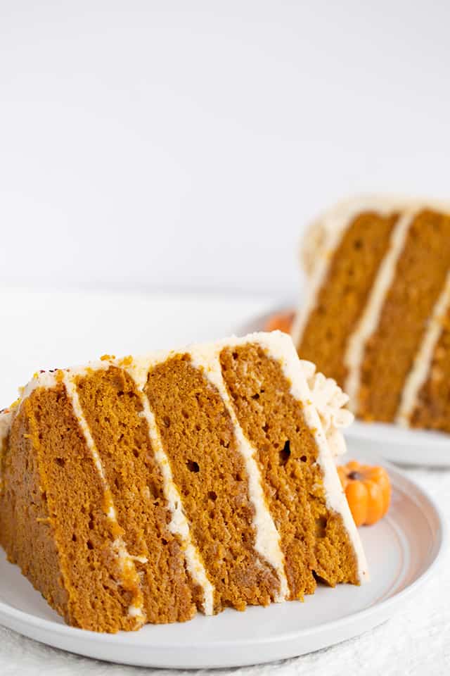 Pumpkin Cake Recipe - This moist Pumpkin Spice Cake with Brown Butter Frosting is one of the tastiest fall desserts! The brown butter frosting has specks in it and tastes sweet and nutty. The pumpkin cake is packed full of spices and topped with adorable chocolate pumpkin truffles. #pumpkin #cake #falldesserts #cookiedoughandovenmitt 2 slices of pumpkin cake on their sides on white dessert plates