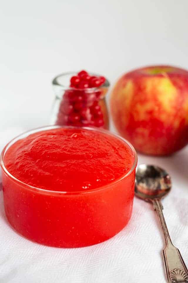 angled shot of glass of red hot applesauce showing the top of the applesauce, candies, apple, and spoon