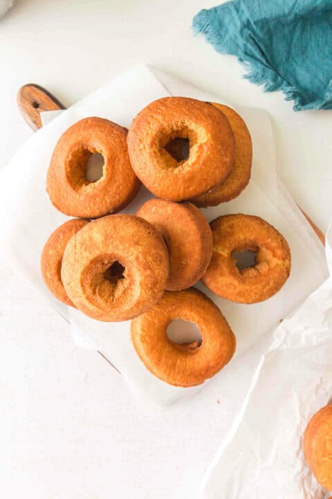 A pile of plain, golden-brown homemade cake donuts arranged on a piece of parchment paper on a white surface.