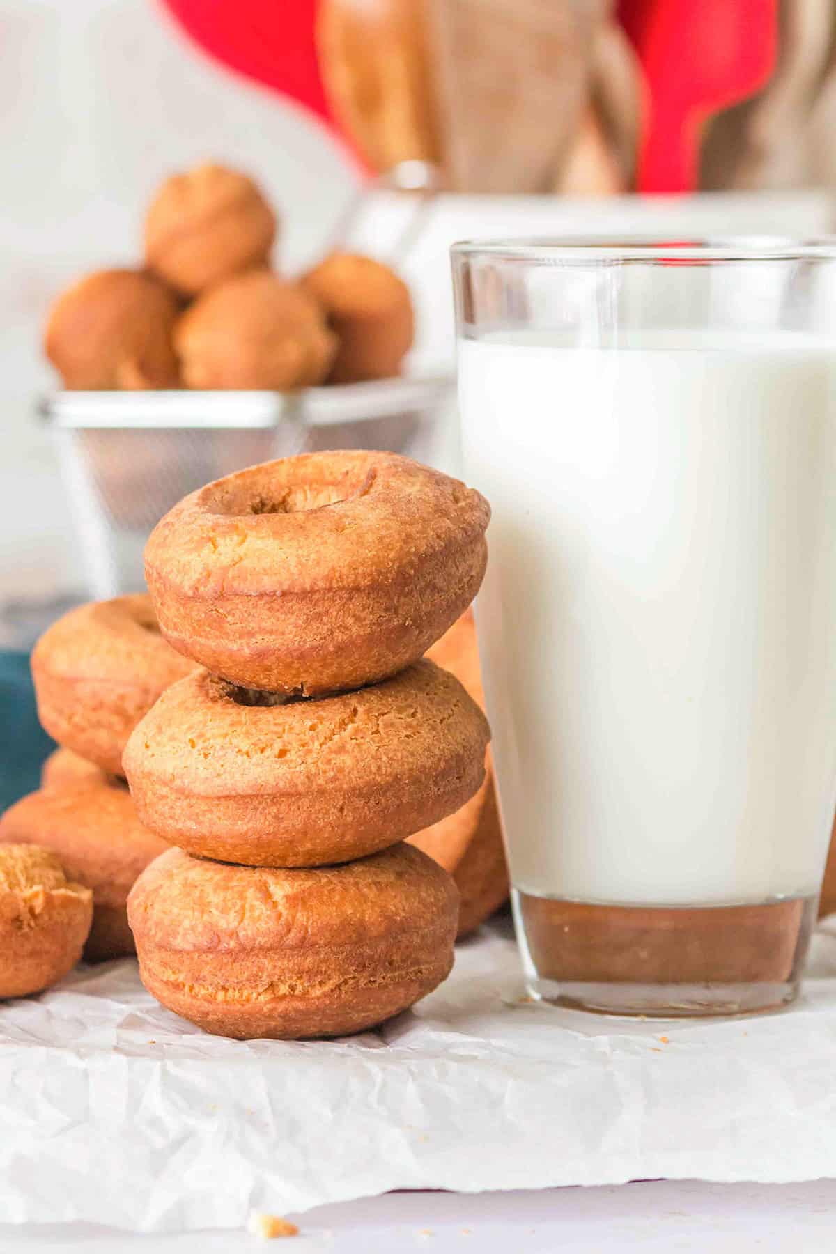 A stack of three homemade cake donuts sits next to a glass of milk, with more donuts in the background on a crumpled white surface.