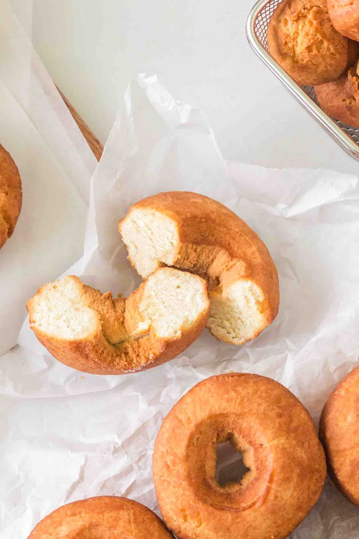A broken homemade cake donut rests on parchment paper among other whole donuts, with a tray of more donuts in the corner.