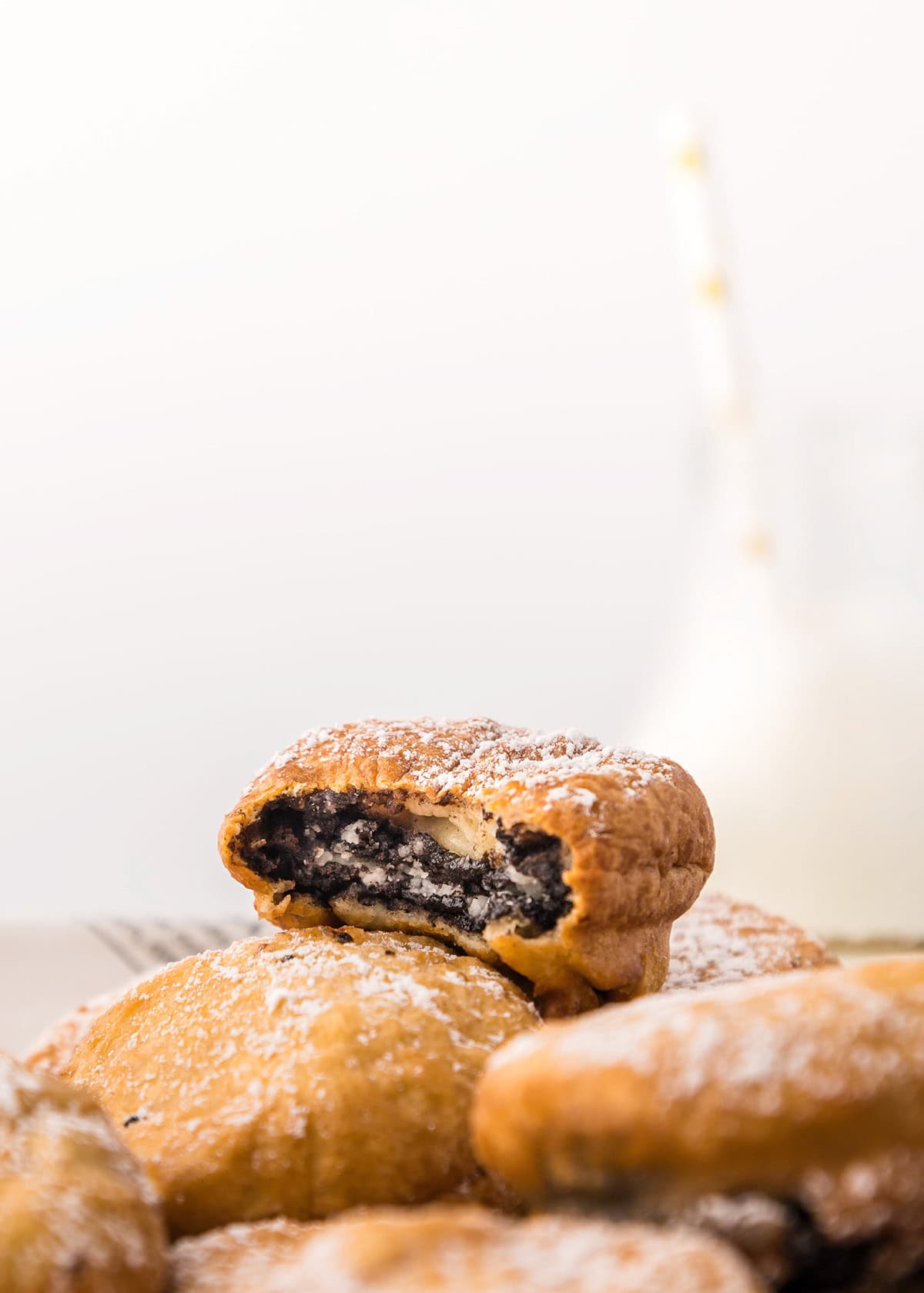 A close-up of deep fried Oreos dusted with powdered sugar; one is bitten to reveal the creamy center. A blurred glass of milk sits in the background.
