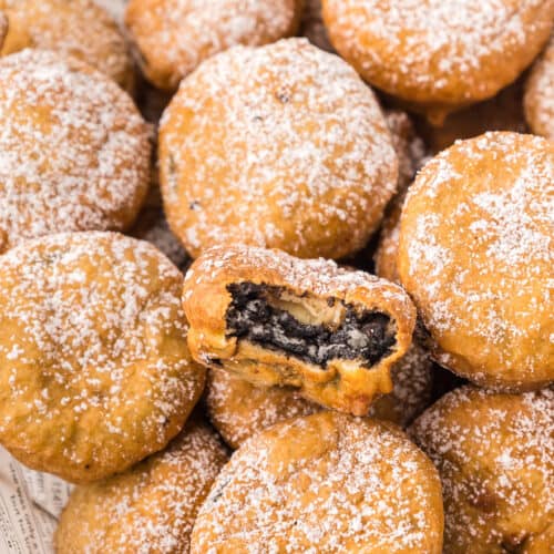A pile of deep fried Oreos dusted with powdered sugar, with one cookie in the center revealing its dark Oreo filling from a bite taken out.