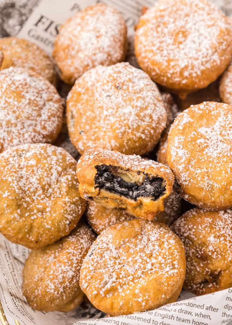 A pile of deep fried Oreos dusted with powdered sugar, with one cookie in the center revealing its dark Oreo filling from a bite taken out.
