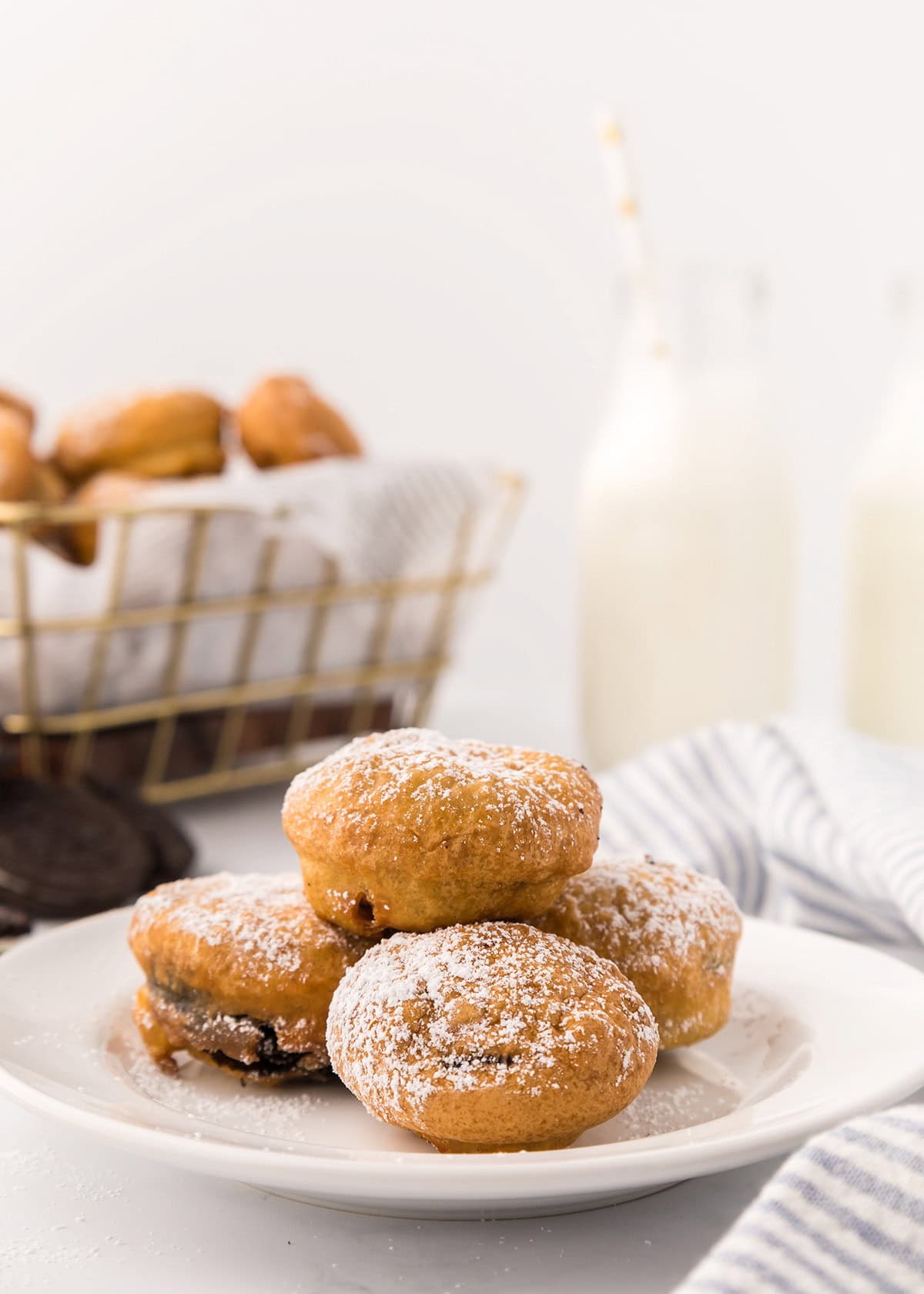 A plate of powdered sugar-dusted deep fried Oreos sits in the foreground, with a wire basket of more treats and two bottles of milk in the background.