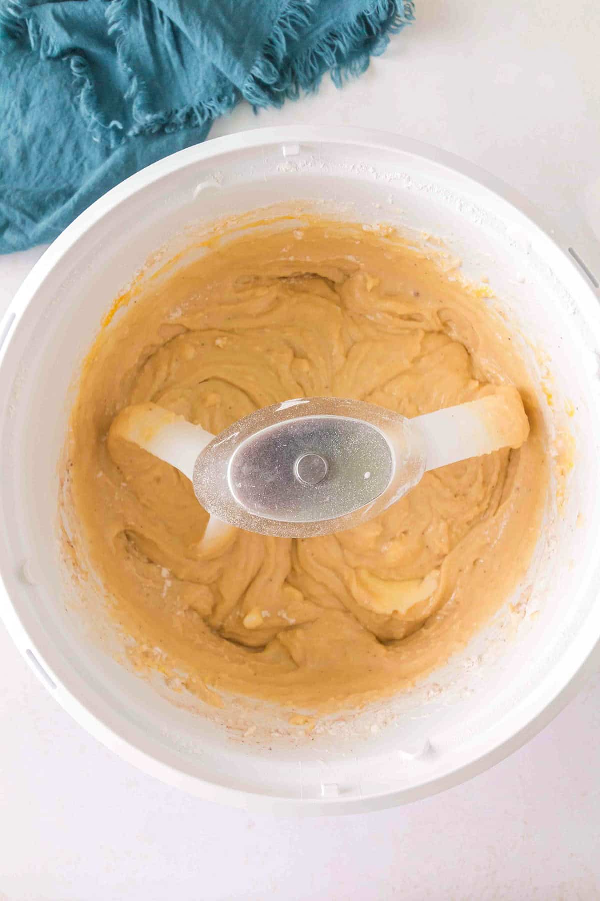 A mixing bowl with thick, light brown batter for Homemade Cake Donuts and a plastic mixing blade, placed on a white surface next to a blue cloth.
