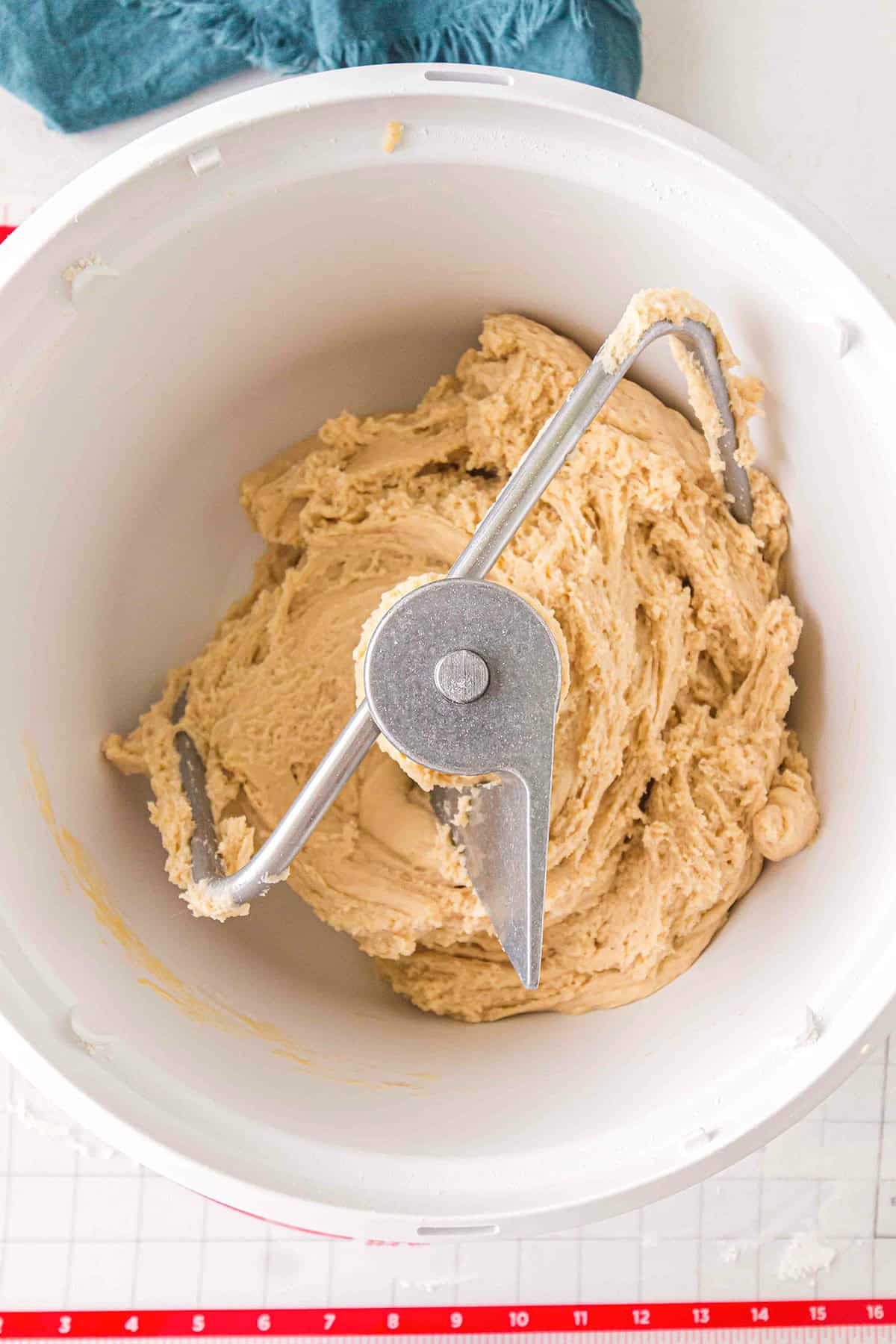 Thick cookie dough mixture in a white mixing bowl with a metal dough hook attachment, viewed from above—perfect for crafting homemade cake donuts.