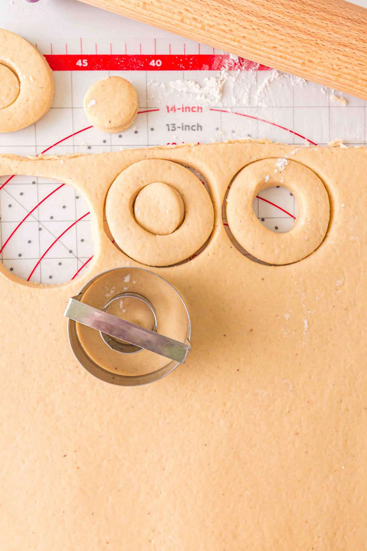 Homemade Cake Donuts are taking shape as round doughnut rings are cut from soft dough with a metal cutter on a floured surface, surrounded by a rolling pin and measurement mat.