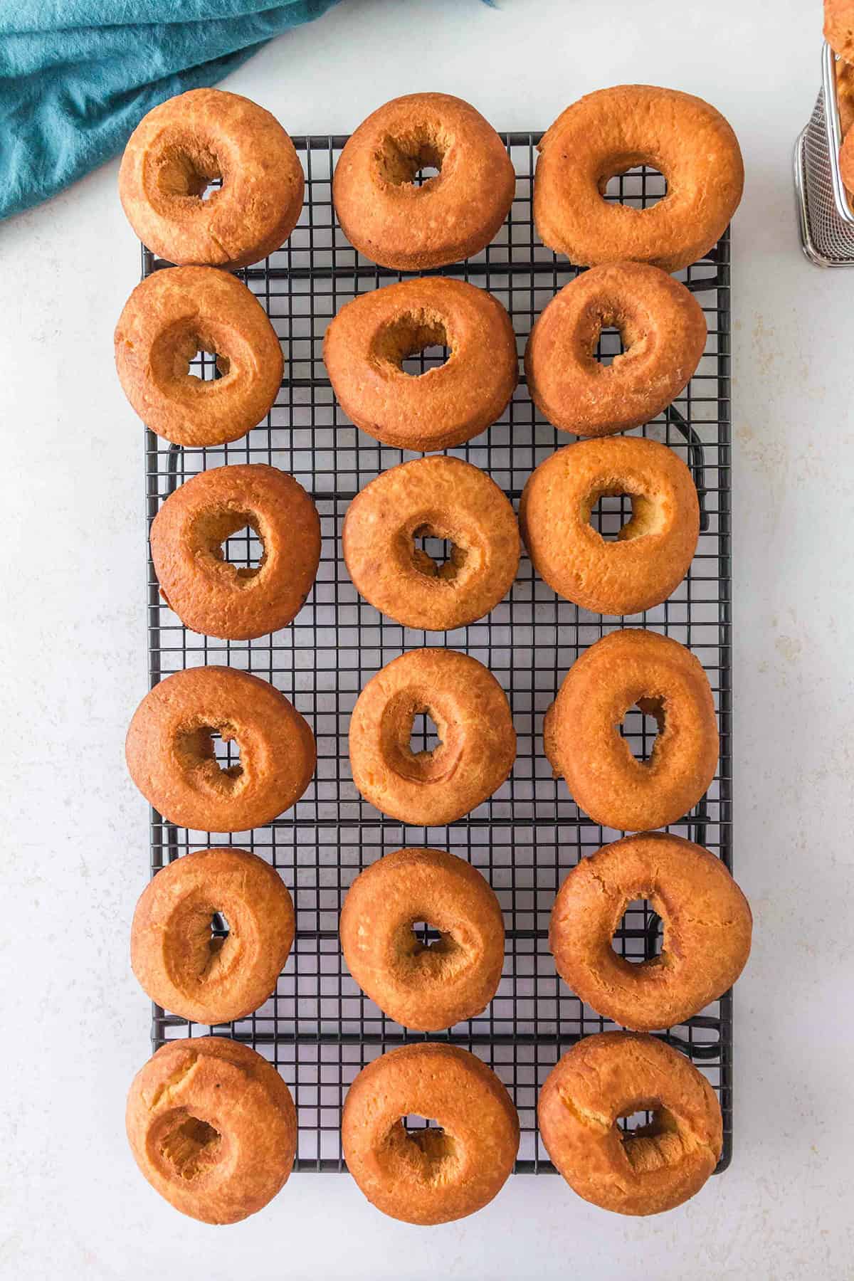 Eighteen plain bagels cooling on a black wire rack, arranged in even rows on a light surface with a blue cloth partially visible in the top left corner—creating a cozy atmosphere that evokes the warmth of baking homemade cake donuts.