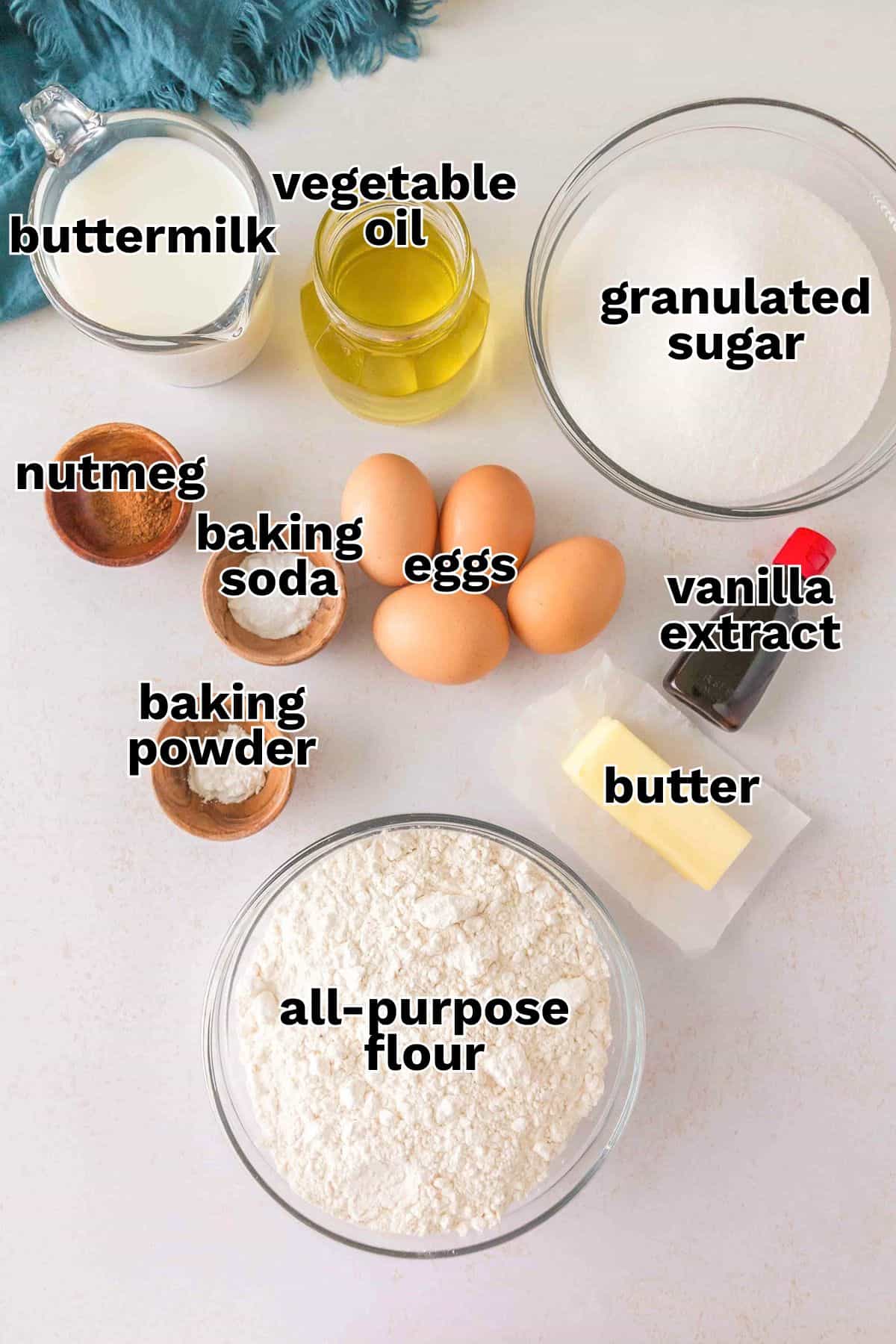 Baking ingredients on a countertop for homemade cake donuts: bowls of flour, sugar, buttermilk, eggs, butter, vegetable oil, plus vanilla extract, baking soda, baking powder, and nutmeg in small containers.