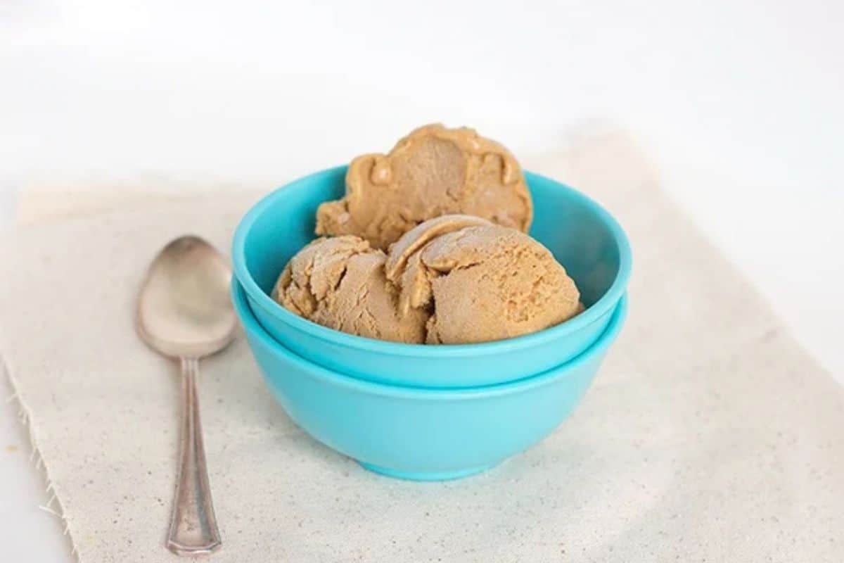Three scoops of tan pumpkin pie ice cream in a light blue bowl next to a metal spoon.