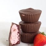 Three stacked Strawberry Buttercream Cups are shown next to a cut-open chocolate cup revealing a pink filling. A whole strawberry lies beside them on a light surface.