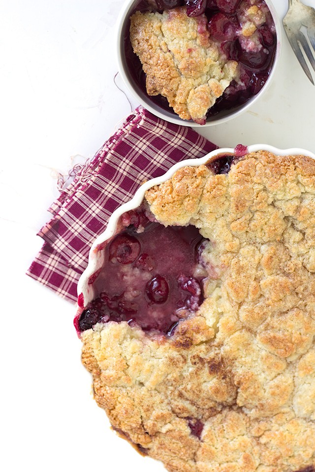 overhead image of a fresh cherry cobbler with sugar cookie topping. One serving sits in a white ramekin above it