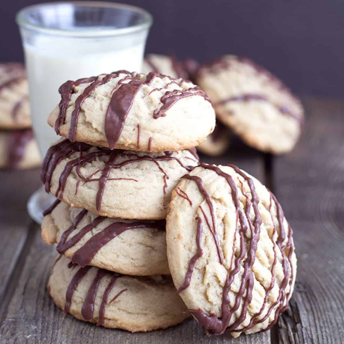 A stack of round coffee cookies with chocolate drizzle sits on a wooden table, with a glass of milk in the background.