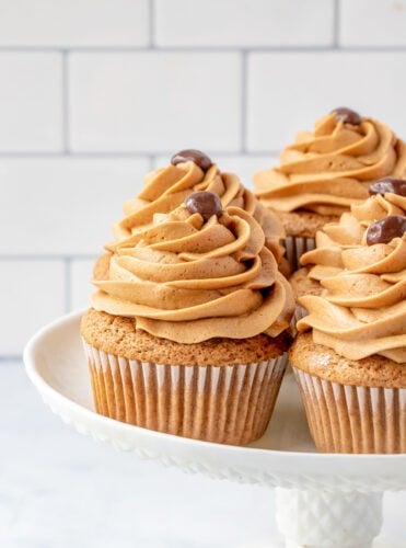 Three coffee cupcakes with swirled brown frosting and chocolate decorations on top, displayed on a white cake stand against a tiled background.