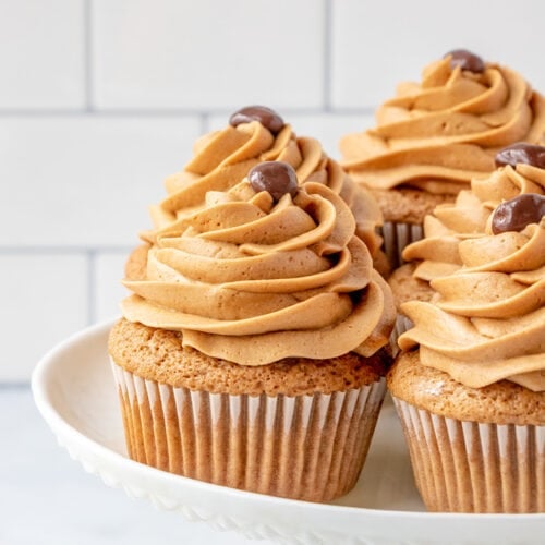 Three coffee cupcakes with swirled brown frosting and chocolate decorations on top, displayed on a white cake stand against a tiled background.