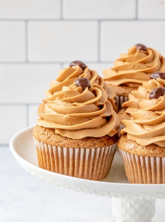 Three coffee cupcakes with swirled brown frosting and chocolate decorations on top, displayed on a white cake stand against a tiled background.