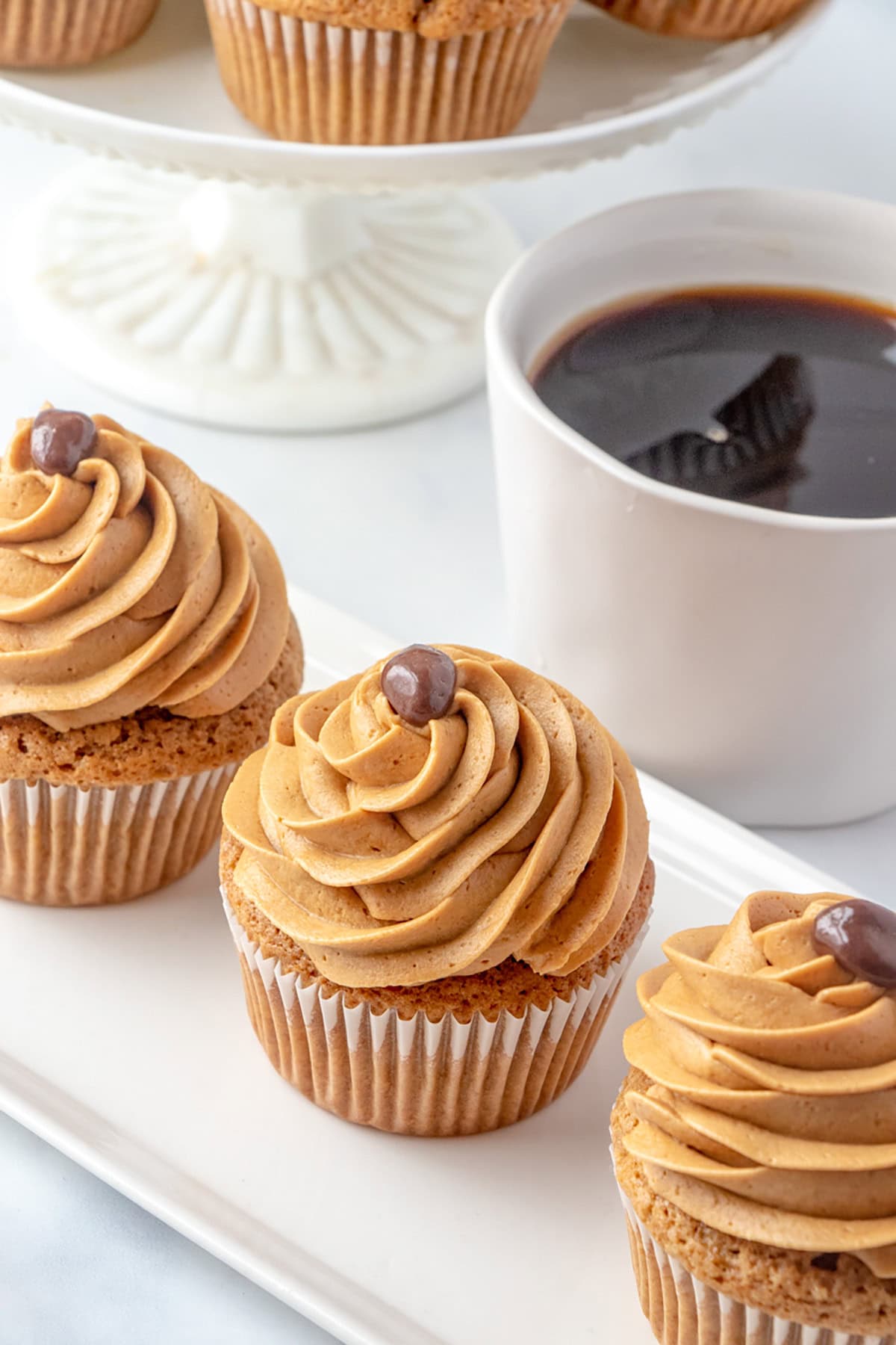 Three Coffee Cupcakes with brown swirled frosting and a small chocolate decoration are arranged on a rectangular plate next to a white mug of black coffee. More cupcakes on a stand are visible in the background.