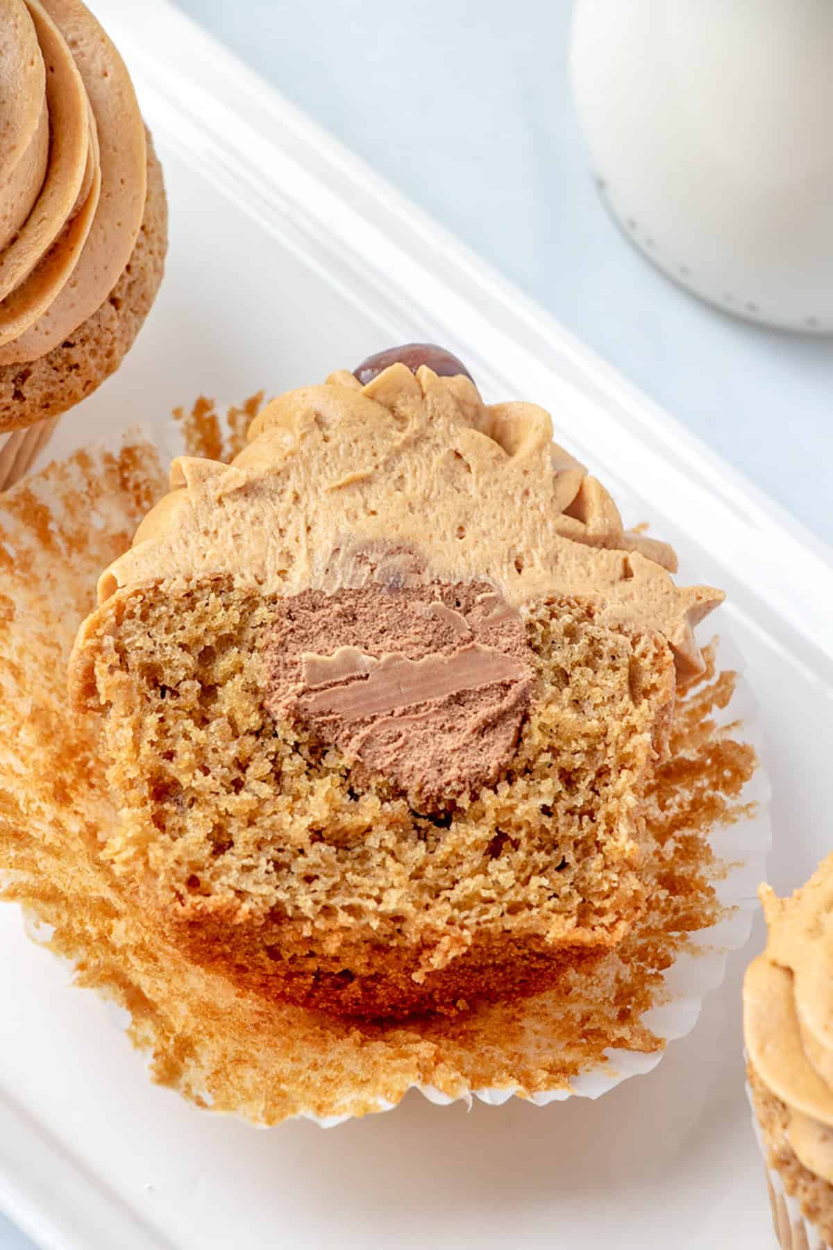 A sliced Coffee Cupcake with brown frosting reveals a chocolate cream filling inside, sitting on a white tray with its wrapper partially peeled.
