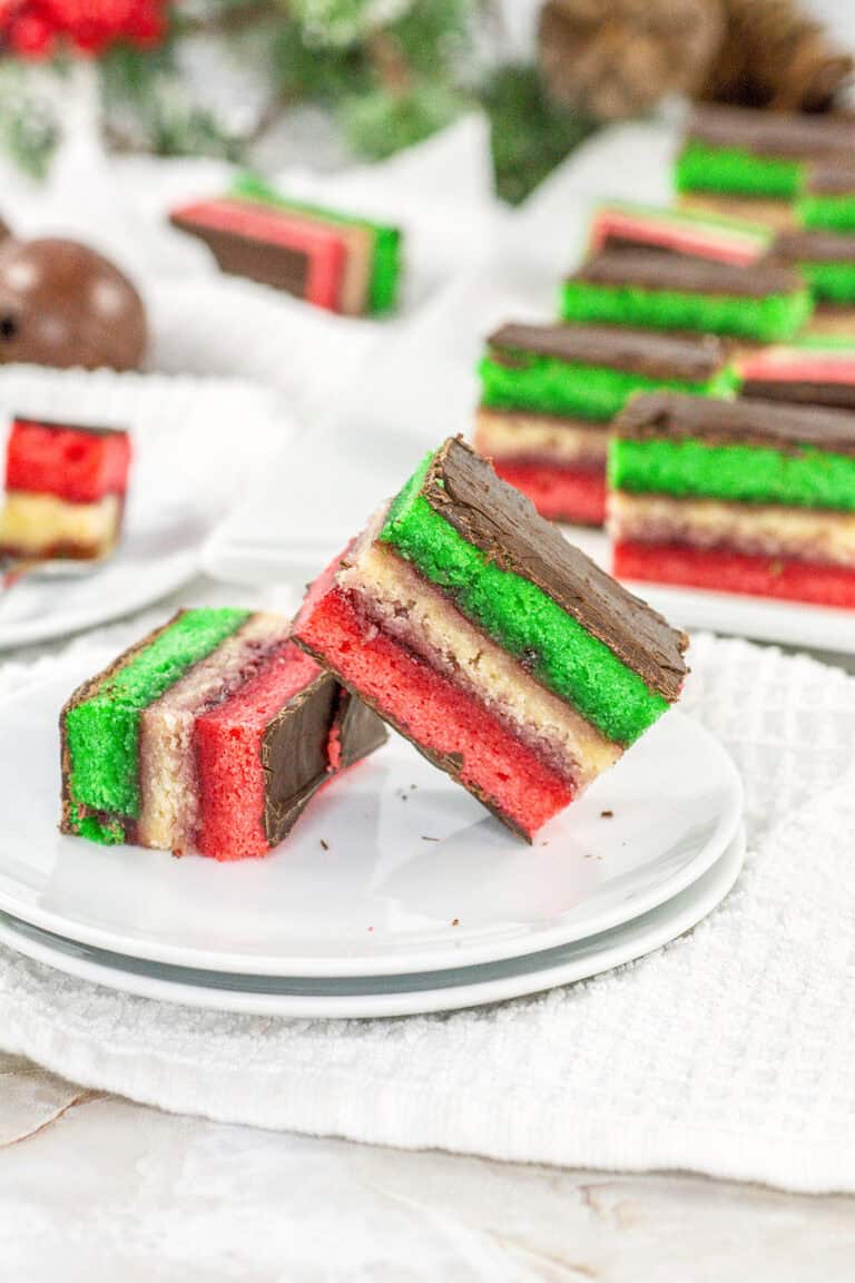 Two slices of layered Rainbow Cookies with red, green, and yellow layers and a chocolate coating are placed on a white plate. More Rainbow Cookies are blurred in the background.