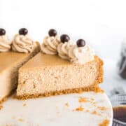 A sliced Coffee Cheesecake with a graham cracker crust, topped with whipped cream swirls and chocolate-covered coffee beans, sits elegantly on a marble cake stand.