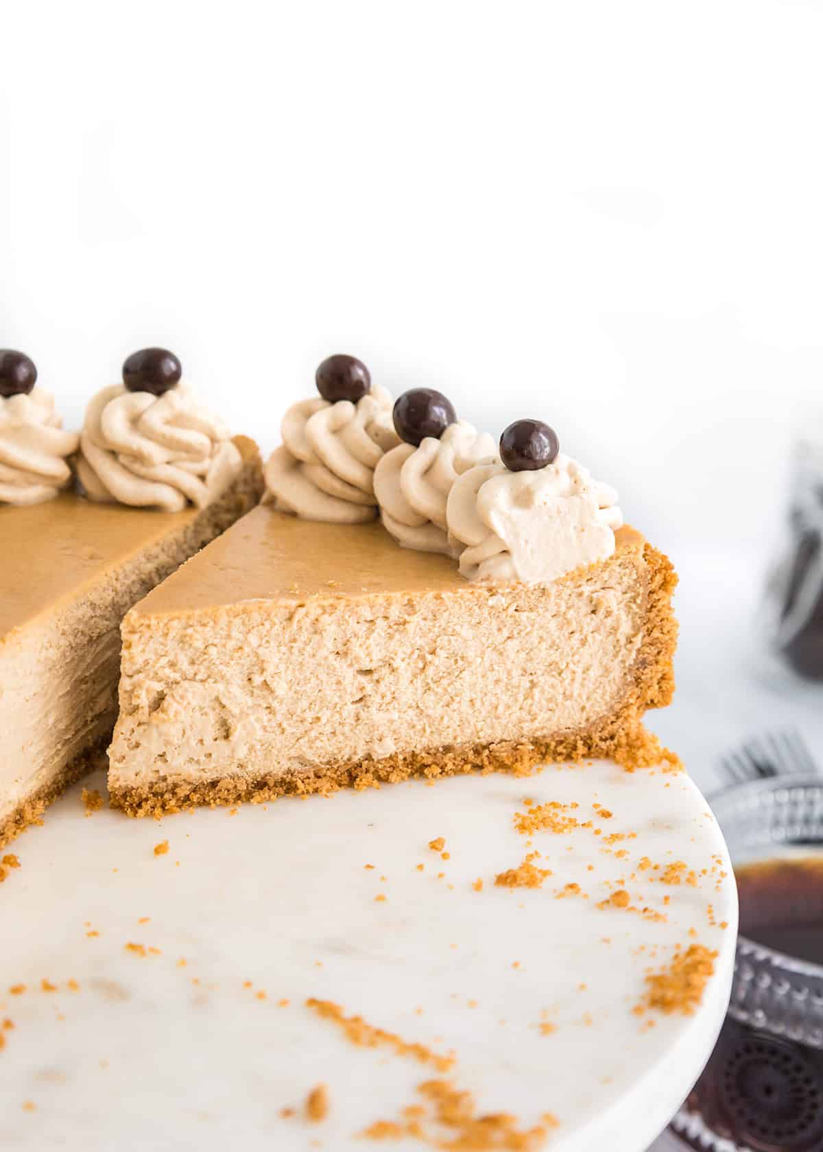 A sliced Coffee Cheesecake with a graham cracker crust, topped with whipped cream swirls and chocolate-covered coffee beans, sits elegantly on a marble cake stand.