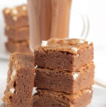 stack of hot cocoa brownies on a white square plate with a mug of hot cocoa behind it