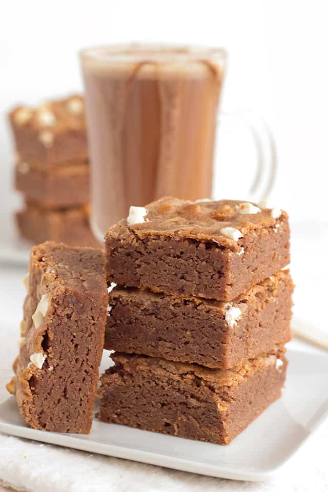 stack of hot cocoa brownies on a white square plate with a mug of hot cocoa behind it