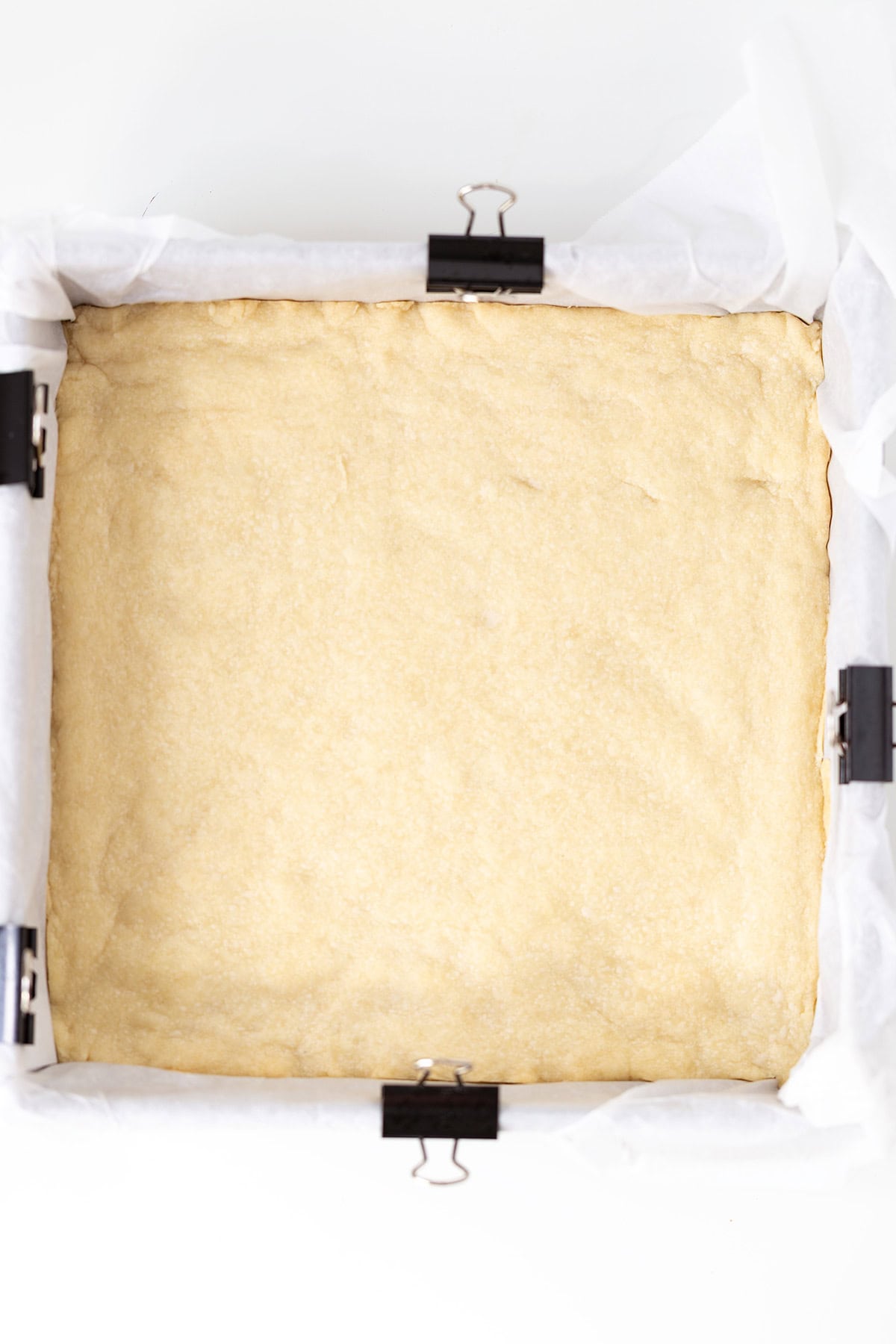 A square baking pan lined with parchment paper and clips holds an even layer of unbaked dough, ready to be transformed into delicious Strawberry Cheesecake Bars.