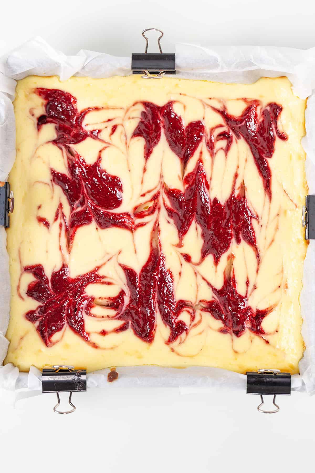 A square pan lined with parchment paper and clips holds beautiful Strawberry Cheesecake Bars, featuring a vibrant red raspberry swirl pattern on top.