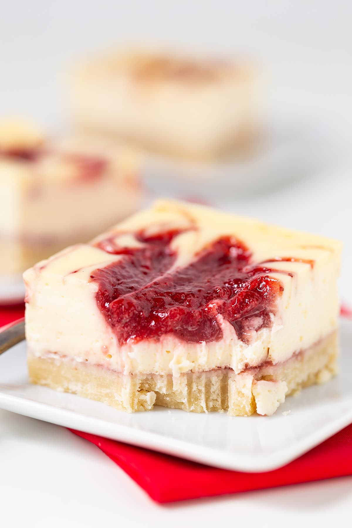 A close-up of Strawberry Cheesecake Bars with a swirl of red berry sauce on top, served on a white plate with a bite taken out of the corner.