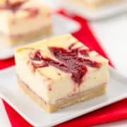A square slice of Strawberry Cheesecake Bar with a swirl of red berry sauce on top, served on a white plate with a red napkin underneath.
