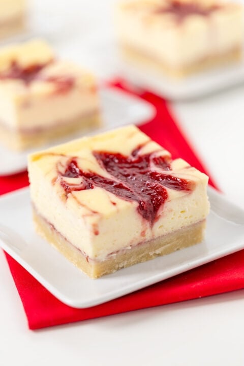 A square slice of Strawberry Cheesecake Bar with a swirl of red berry sauce on top, served on a white plate with a red napkin underneath.