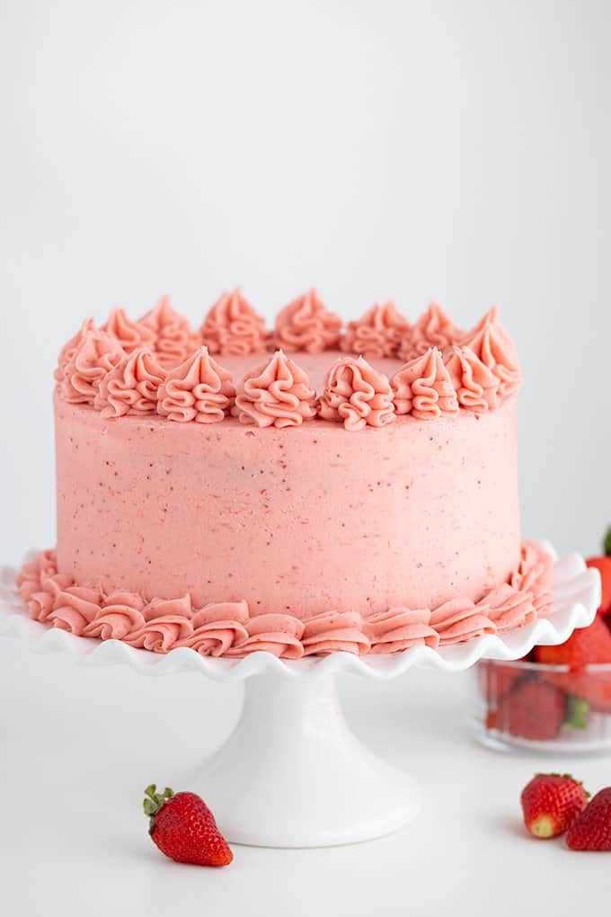 whole strawberry cake on a white cake plate with strawberries under it on a white surface