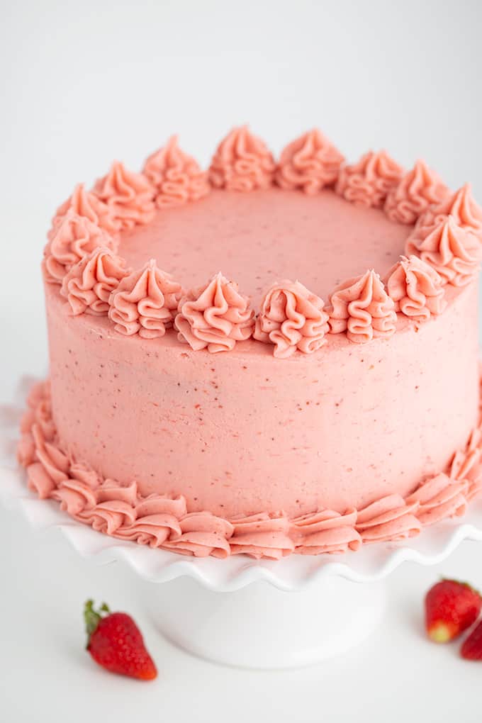 whole homemade strawberry cake with strawberry frosting on a white cake plate with fresh strawberries scattered around it on a white surface