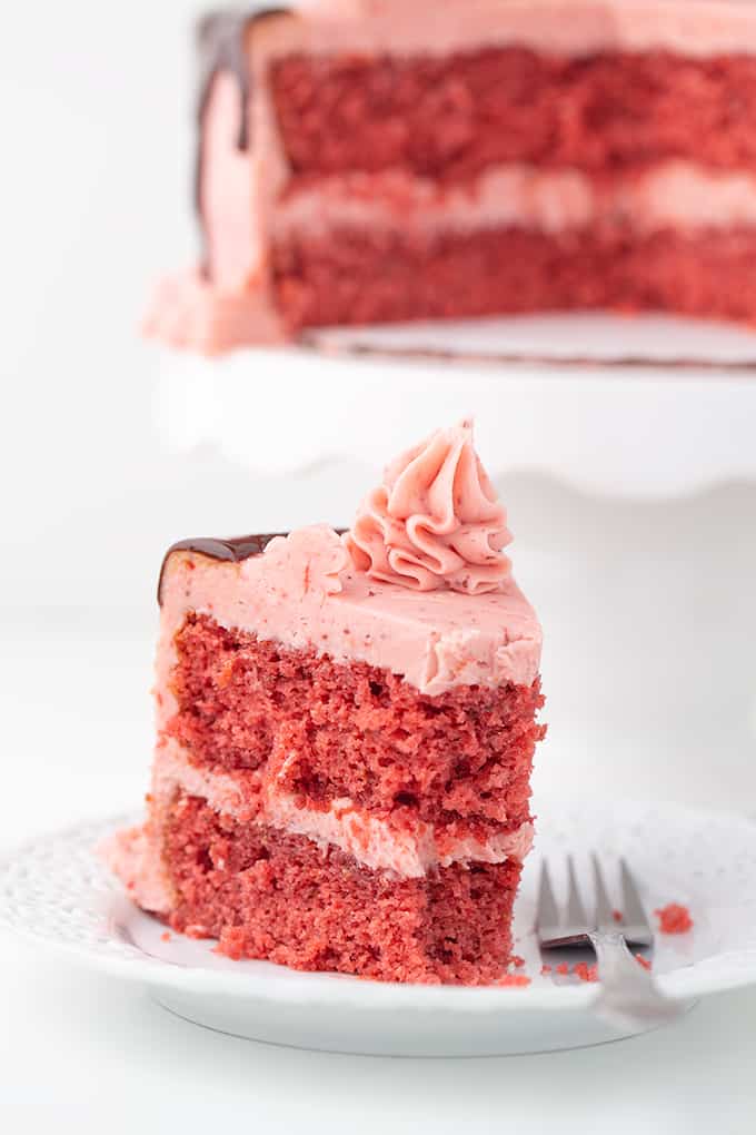 slice of strawberry cake on a white plate with a fork on a white surface with the rest of the cake behind it