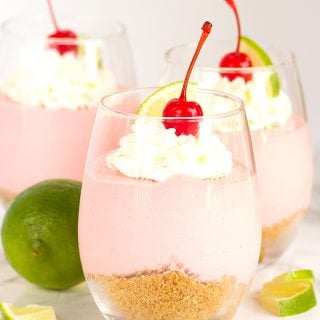 Three pink No Bake Cherry Limeade Cheesecakes served in glasses with whipped cream and a cherry on top.