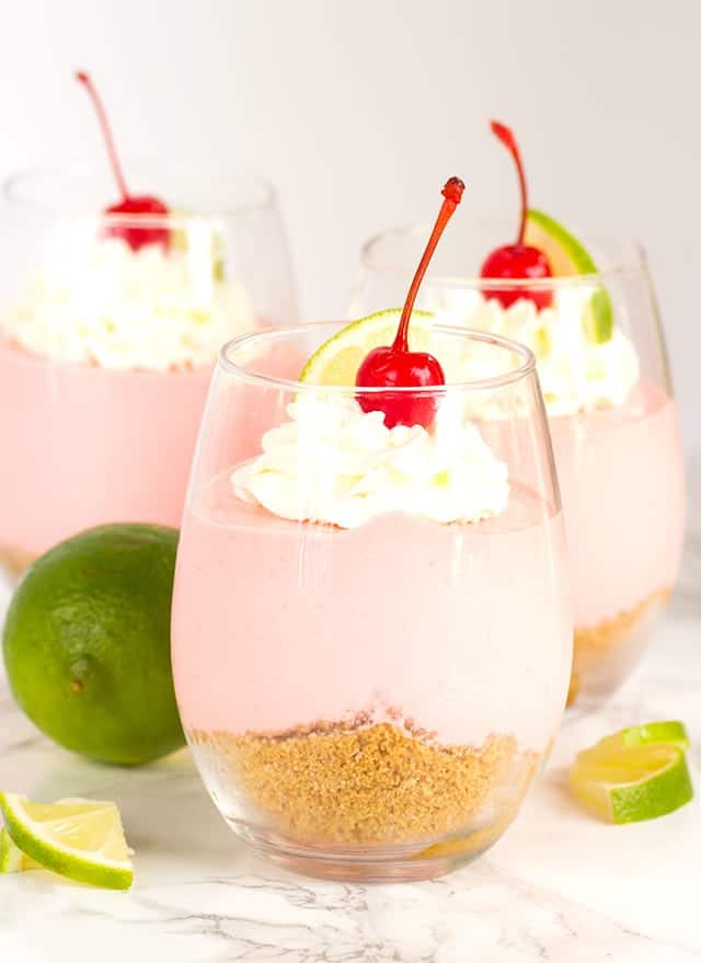 Three pink No Bake Cherry Limeade Cheesecakes served in glasses with whipped cream and a cherry on top.