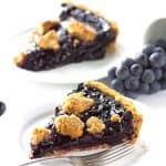 A slice of blueberry pie on a plate with grapes and concord grape tart.