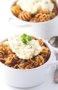 Two white bowls filled with pasta in tomato sauce, reminiscent of a cozy Lasagna Soup Recipe, topped with a dollop of ricotta cheese and a sprig of parsley. A spoon is placed beside one bowl.