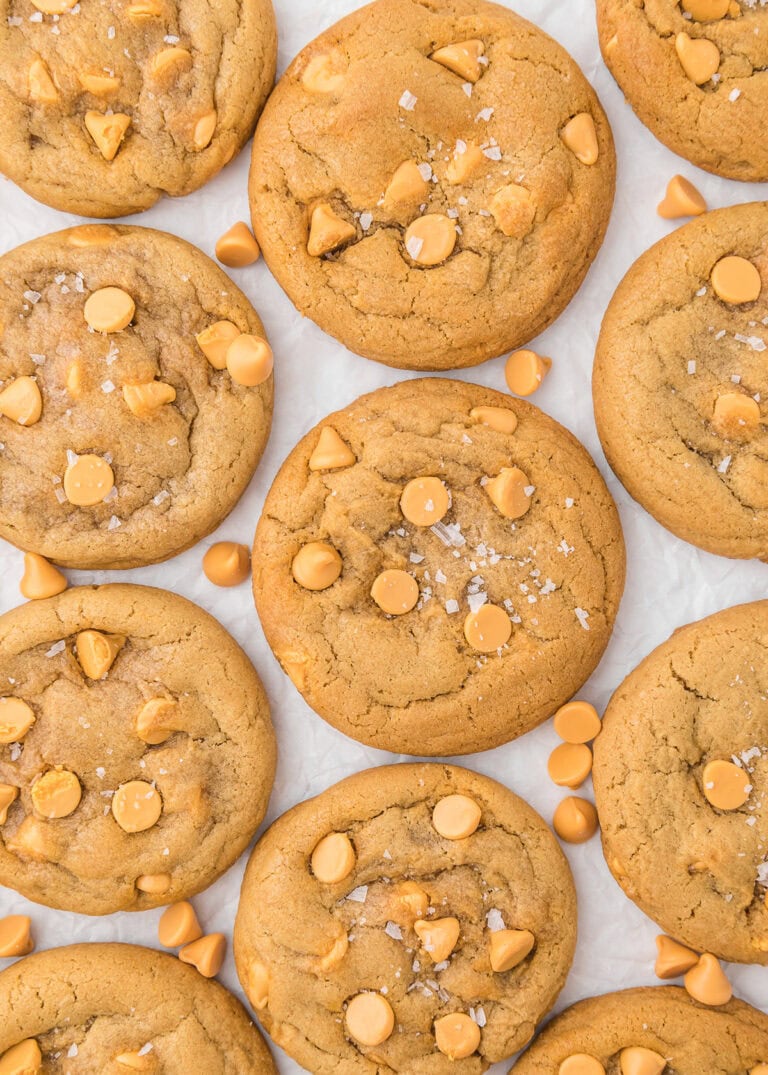 Golden brown butterscotch cookies topped with chips and flakes of sea salt, arranged on a white surface.