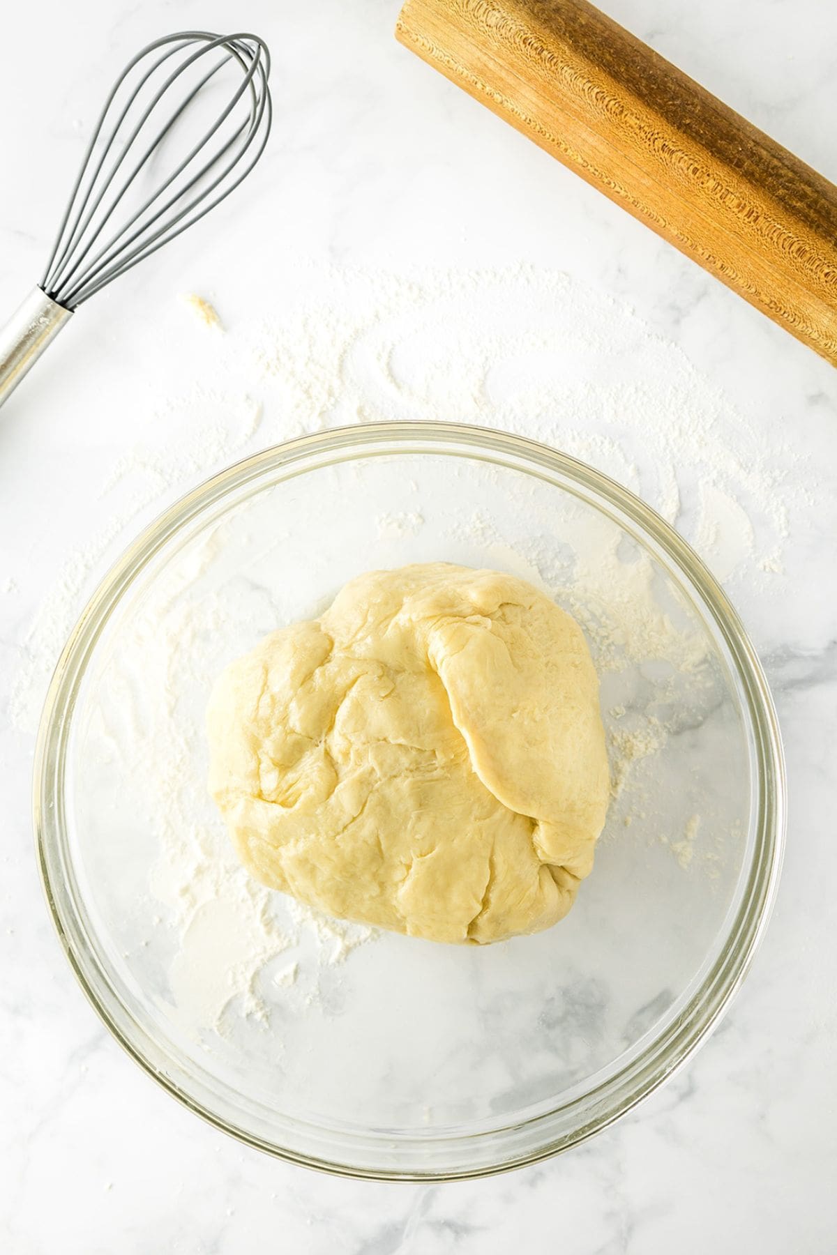 A ball of dough for Potato Donuts sits in a glass bowl on a floured surface, with a whisk and wooden rolling pin nearby.