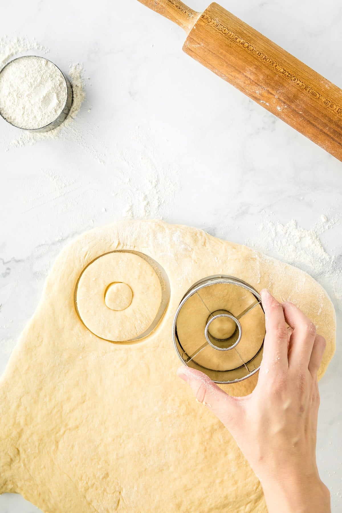 Hand using a round cutter to cut Potato Donuts shapes from rolled dough on a floured surface, with a rolling pin and bowl of flour nearby.