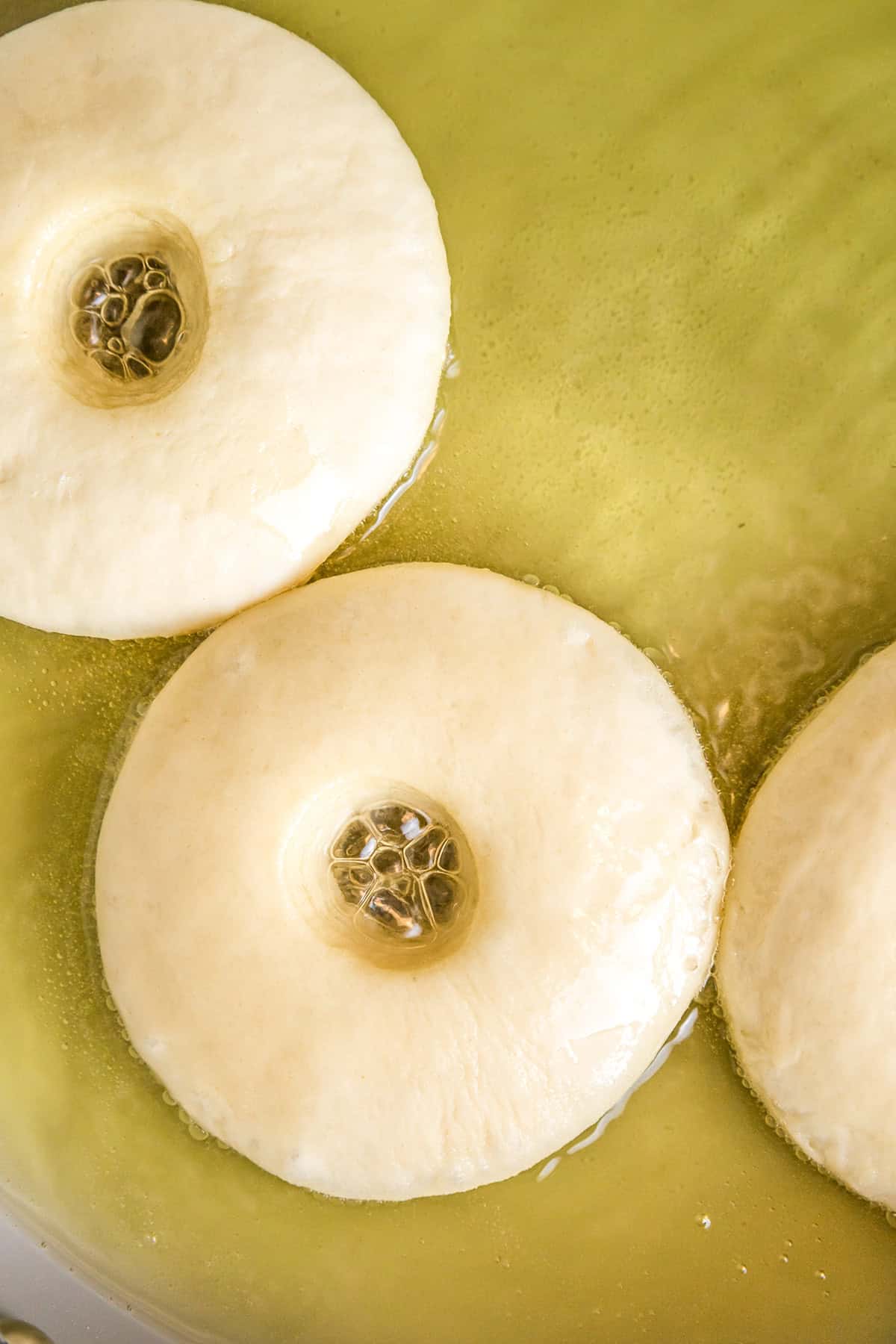 Three round potato donuts with holes in the center are frying in hot oil, partially submerged, with bubbles forming around the edges.