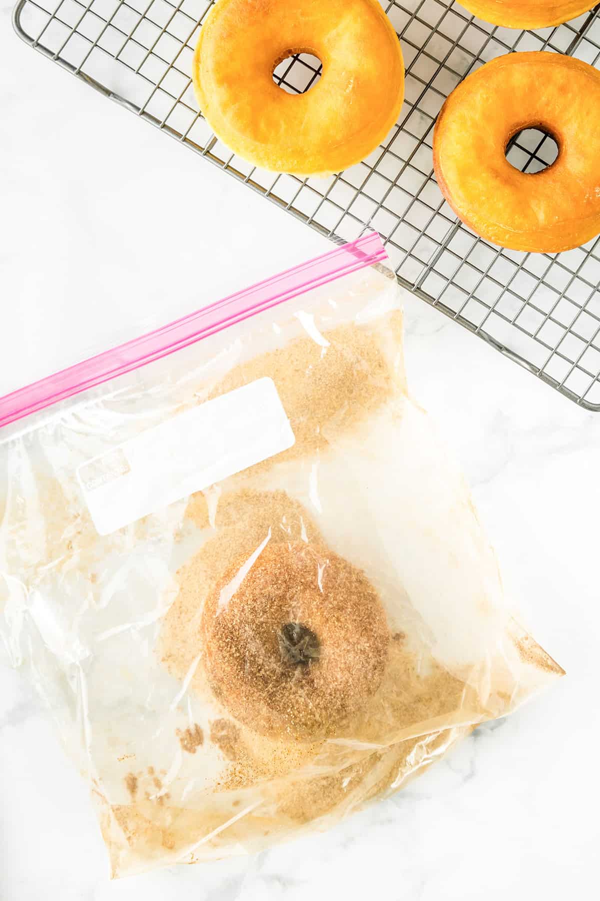 A Potato Donut coated in cinnamon sugar sits inside a zip-top bag, with more potato donuts cooling on a wire rack above on a white countertop.