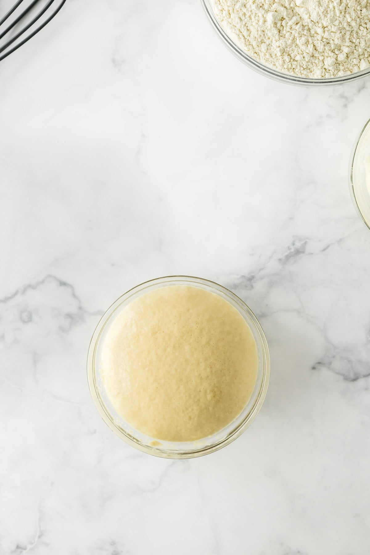 Small glass bowl of active yeast mixture on a marble countertop, with a partial view of a bowl of flour and another ingredient nearby—perfect for preparing homemade Potato Donuts.