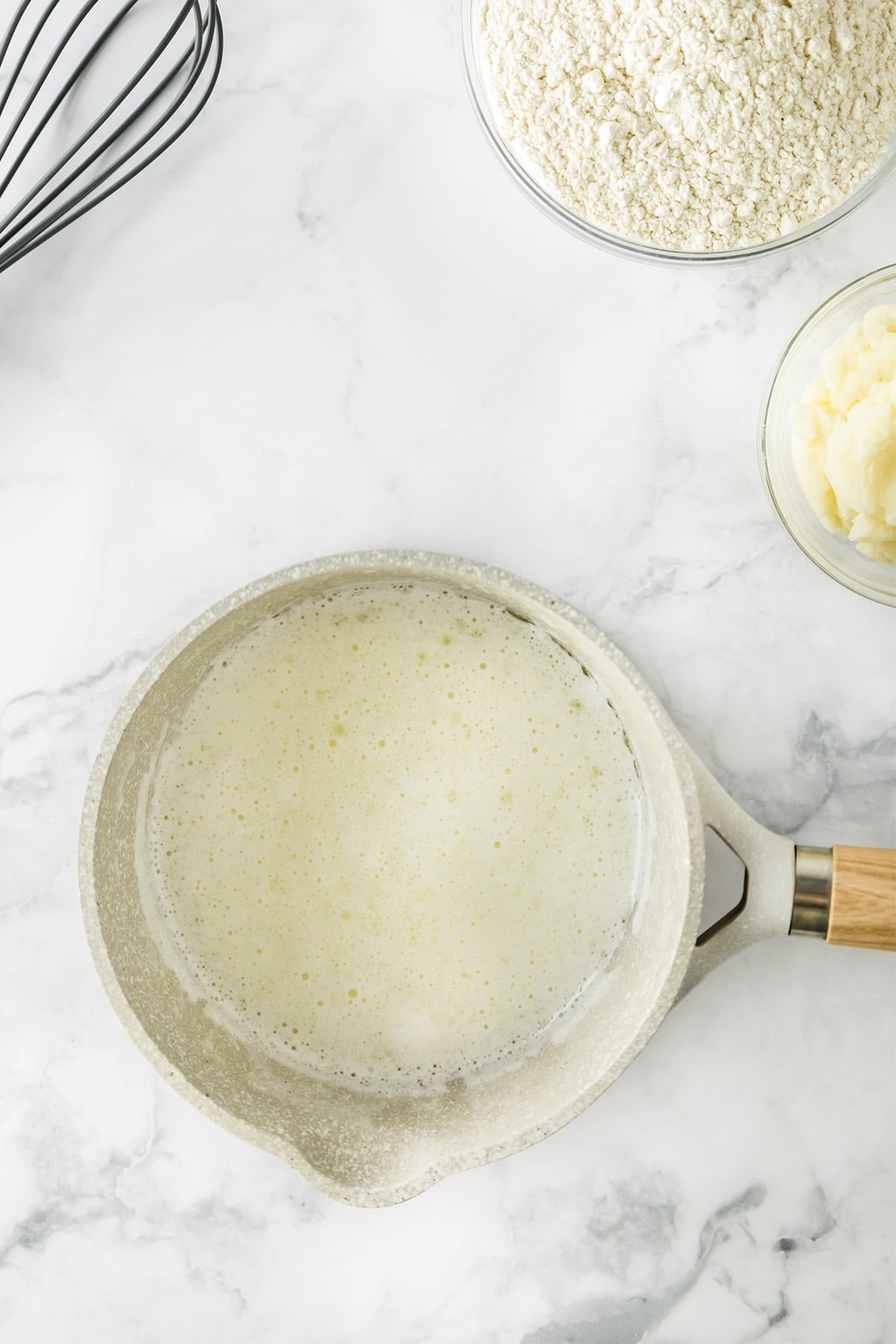 A saucepan with melted butter sits on a marble countertop next to bowls of flour and mashed potatoes, ready for making delicious Potato Donuts, with a whisk nearby.