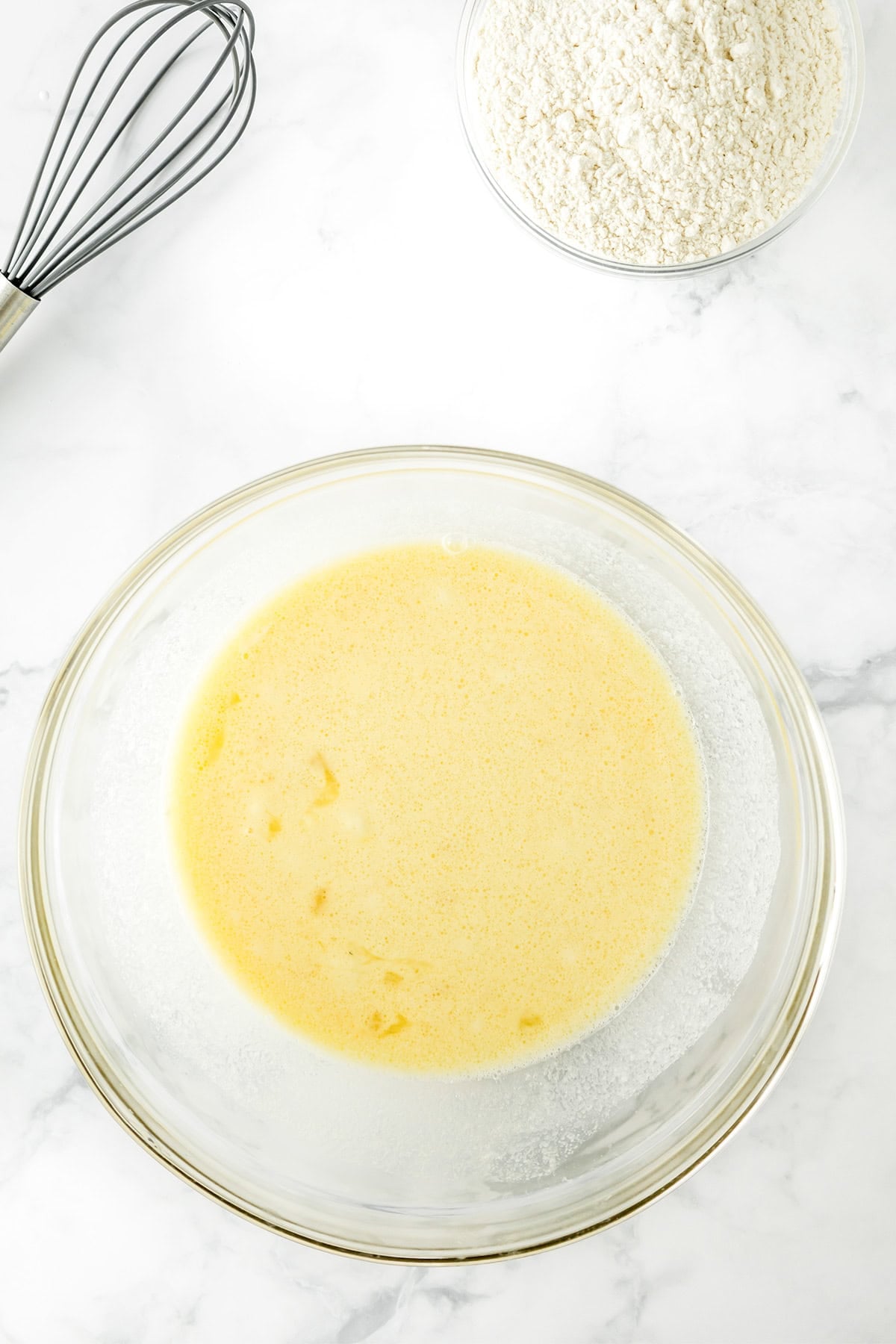 A glass bowl with mixed wet ingredients, a bowl of flour, and a whisk on a white marble surface are ready for making delicious Potato Donuts.