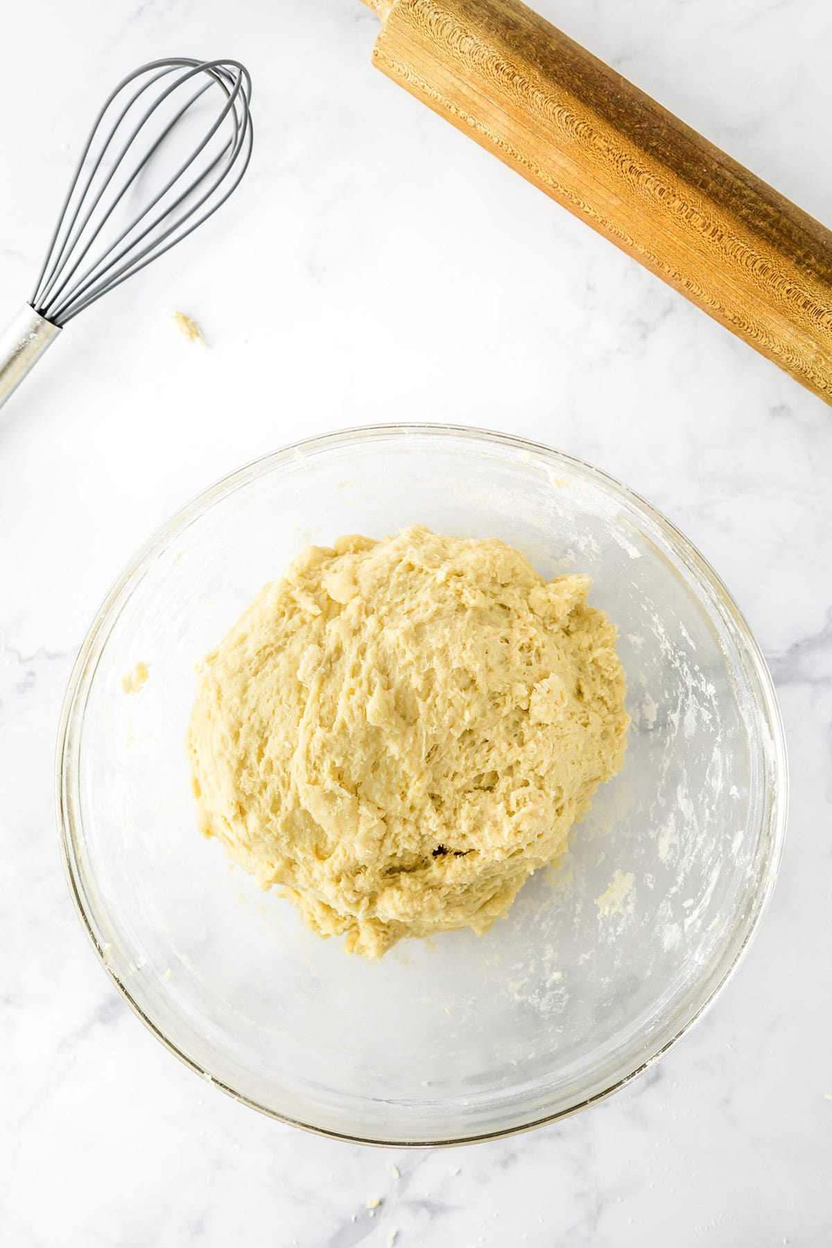Ball of dough in a glass bowl on a marble surface, with a metal whisk and wooden rolling pin nearby, ready to be transformed into delicious potato donuts.
